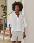 A woman with short curly hair wears the Frank & Eileen ROSIE SS Button Up F25 and light beige shorts, standing indoors in a modern, sunlit room with wooden stairs and plants in the background.