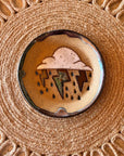 The Monsoon Trinket Dish by Faire features a round ceramic design with a cloud, raindrops, and metallic lightning bolt, displayed on a woven textured placemat—ideal for fans of unique handmade desert decor.