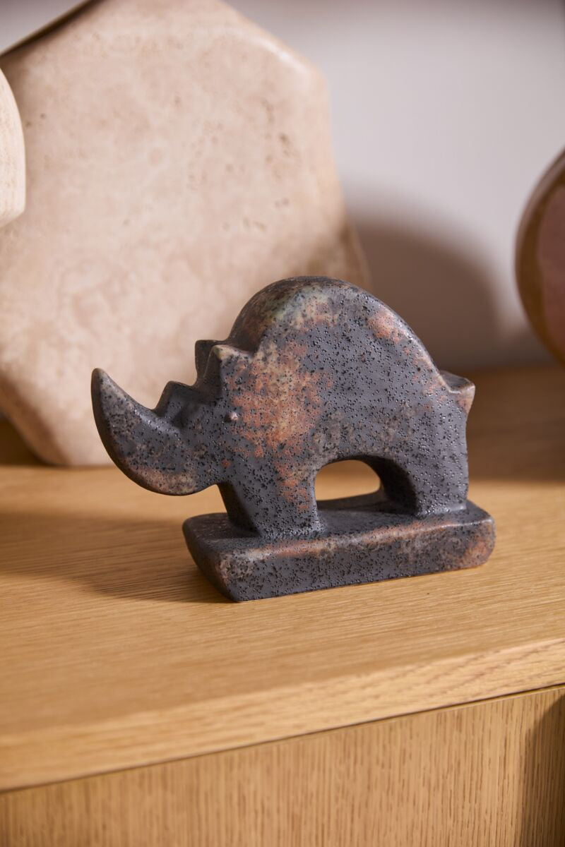 The Accent Decor Rhino Sculpture features a small, dark-speckled design with a curved back and large horn, making it a striking accent on any wooden surface alongside neutral decor.