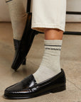 Someone wearing cream pants, black loafers, and Perfectwhitetee's You Are Perfect Sock—a vintage-inspired light gray ribbed cotton style with "you are perfect." in black text—sits cross-legged, showing off the visible wording on their sock.