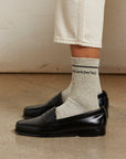 A person stands on a concrete floor against a light wall, wearing light beige pants, Perfectwhitetee's You Are Perfect Sock (vintage-inspired ribbed cotton socks with "you are perfect"), and black loafers.