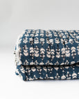 The Faire Blossom Block Print Quilt in navy blue, featuring a white floral pattern, is folded and stacked neatly on a white surface—an ideal bohemian bedding accent.