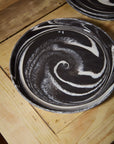 The Marbled Charger by Accent Decor, featuring a handcrafted black stoneware surface with swirling white patterns, sits on a wooden table. Another decorative platter is partially visible in the background.