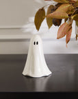 The Accent Decor ghostly Figurine Small is a hand-painted, white ceramic ghost with black eyes, perfect for charming Halloween décor. It sits on a dark surface with autumn leaves hanging in the top right corner of the image.