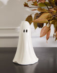 The Accent Decor Ghostly Figurine Large, a white ceramic ghost with black eye holes, is displayed on a black surface beside autumn leaves in brown, orange, and yellow—ideal for Halloween décor.