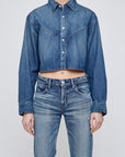 A person wears the Moussy Foley Shirt, a women's cotton denim cropped button-down jacket with long sleeves and a collar, paired with high-waisted blue jeans, standing against a plain light background.