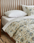 A bed is styled with Coyuchi Inc's Flora Org Print Duvet Undyed Lagoon King/CKing, showcasing a blue and yellow botanical design, beside a wooden nightstand holding a lamp, flowers in a vase, and a book on top of a woven rug.