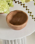 The Cyrus Bowl by Accent Decor, a small round terracotta piece, rests on a white textured surface with green and yellow flowers and leafy stems partially visible around it.