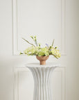 A minimalist arrangement of pale yellow and white blooms in the Cyrus Compote by Accent Decor sits on a white fluted pedestal against a white paneled wall.