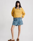 A woman with straight dark hair and glasses poses against a plain background in a Denimist Boxy Cable Sweater, layered over a striped shirt, denim shorts with large front pockets, and brown boat shoes.