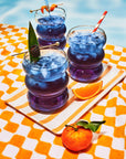 Three Faire Blackberry Jasmine Iced Teas sit in round glasses on a striped tray by a pool, garnished with fruit and striped straws. An orange slice and tangerine rest on an orange-and-white towel nearby.