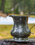 The Vintage Pitcher with Deer by Faire, featuring engraved designs and crafted in Italy, is displayed on a log outdoors with blurred grass, water, trees, and a deer in the background.