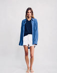 A woman with shoulder-length brown hair stands against a plain background wearing the Denimist SUTTON BUTTON FRONT BRIGHT BLUE shirt over a black V-neck top, paired with white frayed denim shorts and white sandals.