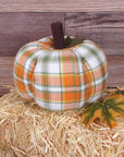 The Faire Pumpkin is a fabric plaid fall decor piece, ideal as a farmhouse ornament. It sits on a hay bale with two artificial autumn leaves, set against a wooden plank background.