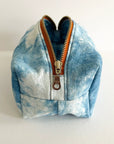 The Faire Dyed Toiletries Bag is a small, box-shaped pouch with natural blue and white tie-dye, brown zipper trim, and a silver pull, shown on a plain white background.