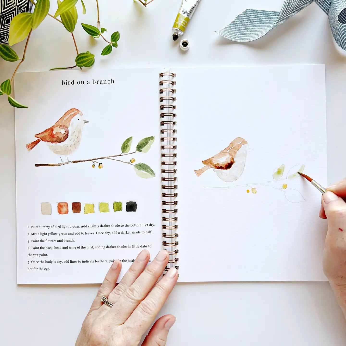 In their spiral-bound sketchbook, a person paints a watercolor bird on a branch with beginner-friendly steps, color swatches, and text instructions. Art supplies and a plant rest beside them, evoking pages from Faire's Watercolor Workbook.