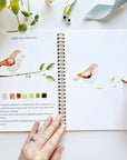 In their spiral-bound sketchbook, a person paints a watercolor bird on a branch with beginner-friendly steps, color swatches, and text instructions. Art supplies and a plant rest beside them, evoking pages from Faire's Watercolor Workbook.