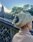 A woman with blonde hair pinned up by Faire’s Hand Painted Pigeon Clip stands near an ornate stone staircase outdoors, framed by trees and a partly cloudy sky.