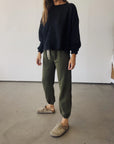 A person wearing a loose black long-sleeve shirt, Hey Gang's The Sweatpants in olive green, and taupe slip-on clogs stands indoors on a concrete floor. Their face is not visible.