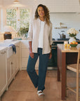 In a bright, modern kitchen, a woman wears Frank & Eileen’s Catherine Favorite Sweatpant with a white button-up shirt as she leans casually against a wooden island surrounded by white cabinetry, plants, and a bowl of fruit.