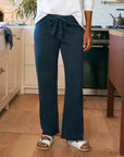 In a kitchen with light cabinets and a wooden floor, a person (shown from shoulders down) wears Frank & Eileen's Catherine Favorite Sweatpant in navy blue, paired with a white long-sleeve shirt and white sandals.