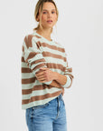 A woman with light hair wears the One Grey Day Sloane Cashmere Boxy sweater, styled as a brown and white striped crewneck pullover with blue jeans, standing against a white background and looking at the camera with folded arms.