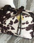 The Faire Oakley Overnighter - Faux Cowhide is a vegan rectangular weekender bag with a dark brown and white cowhide pattern, brown leather handles, and a yellow tag. Handmade in the USA, it's displayed on rustic wood with a weathered backdrop.