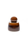 The Faire Amber Stash Jar is a small, hand-thrown ceramic container with a brown glossy amber glaze and rounded lid, shown against a plain white background.