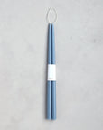 The Floral Society 18" Taper Candle Pair in blue features tall, slender candles bound with a white band and topped with a looped wick—an elegant touch for any home décor. Set on a light gray textured surface.