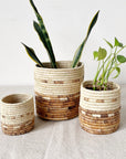 Three Mkongo Plant Baskets by Amsha - Faire, featuring a beige and brown woven pattern, are displayed on fabric; two larger baskets hold green houseplants, while the smallest is empty against a white wall.