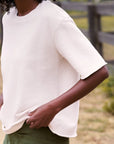 Woman in a casual crew neck tee by Frank & Eileen with green pants stands outdoors near a rustic wooden fence.