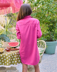 A woman with long brown hair wears the Frank & Eileen MARY Shirtdress S25 in bright pink, standing with her back to the camera beside a polka dot table and lush garden, creating a chic must-have summer vibe.