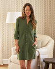 A woman with long brown hair smiles indoors, wearing the Frank & Eileen Southern Dress in green. Her hand is in a pocket. Behind her are a beige armchair, floor lamp, and patterned wallpaper for a relaxed Southern Ireland vibe.