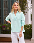 A person confidently smiles outdoors, dressed in a stylish preppy look featuring the Frank & Eileen Patrick Popover Henley and crisp white pants, against a backdrop of lush greenery and large-windowed building.