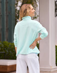 Amidst lush greenery and elegant architecture, a blonde woman stands outdoors, effortlessly chic in Frank & Eileen's Patrick Popover Henley—a preppy light blue 100% cotton turtleneck—and white pants, smiling as she glances to the side.