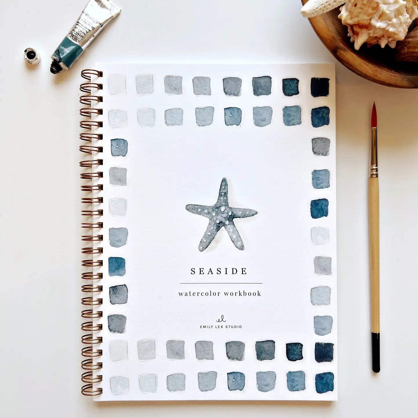The Faire "Watercolor Workbook" features a spiral-bound design with beginner-friendly illustrations. A central starfish is bordered by blue and gray watercolor squares, accompanied by a paint tube, paintbrush, and shell-filled bowl for a serene scene.