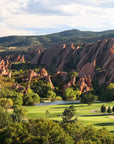 Experience dramatic red rock formations, lush trees, and a top golf course beside a pond—featured in "150 Golf Courses You Need to Play" by Simon & Schuster Inc.—all set against rolling hills and a partly cloudy sky.