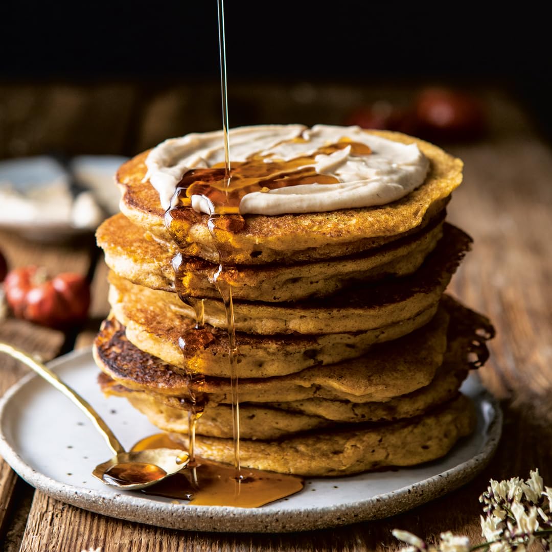 A stack of fluffy pancakes from Half Baked Harvest Super Simple by Random House, topped with creamy spread and syrup, is drizzled with more syrup on a plate with a spoon, set against a rustic wooden background.