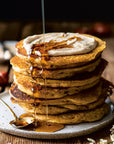 A stack of fluffy pancakes from Half Baked Harvest Super Simple by Random House, topped with creamy spread and syrup, is drizzled with more syrup on a plate with a spoon, set against a rustic wooden background.