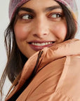 A woman in a colorful Faire Cashmere Beanie and a peach-colored puffer jacket gazes to the side with a gentle smile.