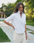 A woman with wavy brown hair stands outdoors on a stone pathway, wearing the Frank & Eileen Patty Popover SS Henley in white and light pants, with greenery and a pool in the background.