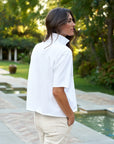 A person with long brown hair, wearing the Frank & Eileen Patty Popover SS Henley in white and light pants, stands outdoors by a pool, looking to the side with lush greenery and trees in the background.