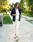 A woman walks outside on a patio, wearing the Frank & Eileen Patty Popover SS Henley in white, cream pants, a dark blazer, and sandals. Lush greenery and a pool are behind her as she glances to the side with a slight smile.
