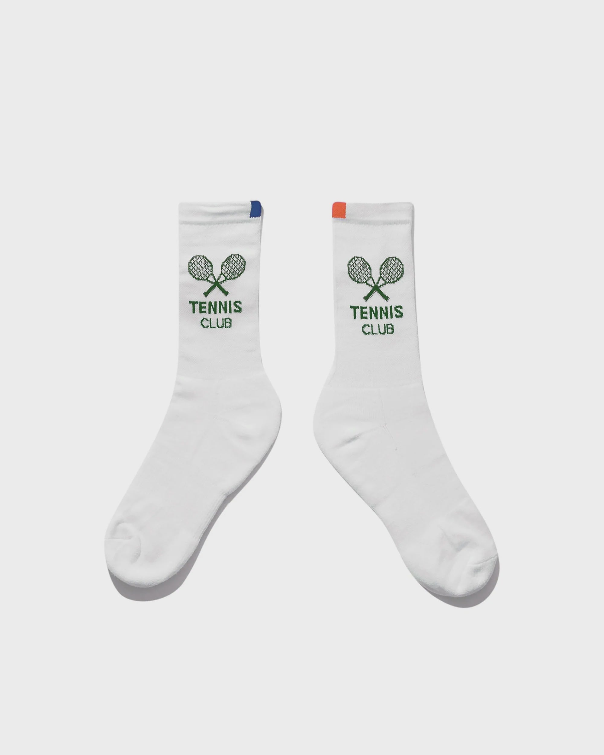 The Women's Tennis Sock WHT by Kule features vintage-inspired white socks with "KULE Tennis Club" and two green tennis rackets. One sock has a blue tab, the other an orange tab at the top for a playful touch.