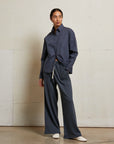 A woman stands indoors against an off-white wall, wearing Perfectwhitetee’s French Terry Pant and a loose dark blue button-up shirt with white shoes. She looks to the side with one hand in her pocket.