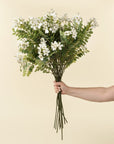 A hand holds the Faire 1-9528 Locust Flower Branch, featuring lush green leafy stems and delicate white blooms, ideal as faux flowers for accent decor against a plain beige background.