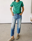 A person poses casually in a green T-shirt called The Crew by Hey Gang, made of 100% cotton, paired with light blue vintage-fit jeans and beige clogs. They stand against a plain white wall on a concrete floor.