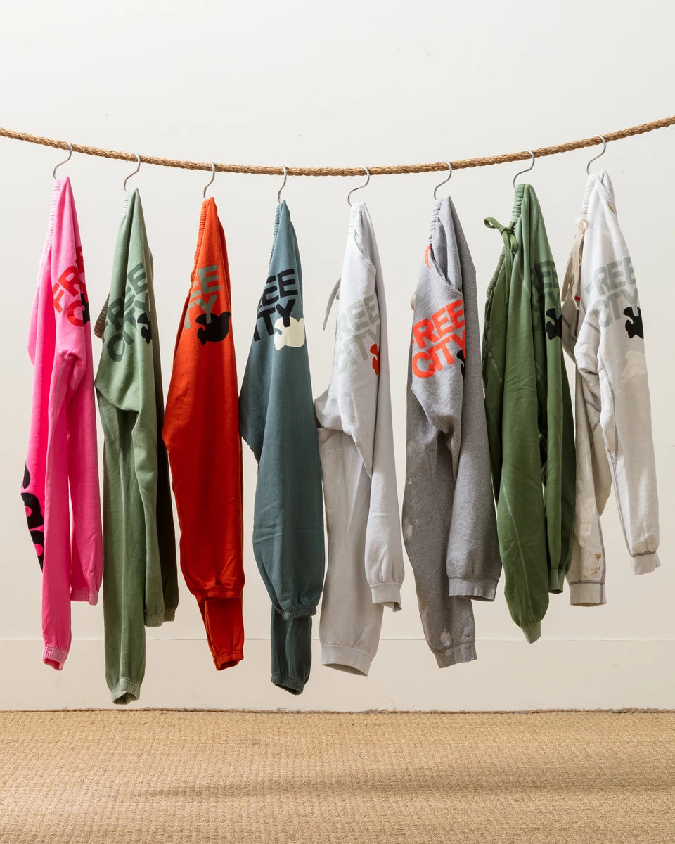 Seven colorful FREECITY sweat pants with various prints hang from a rope against a neutral background, displaying a mix of bright and mute colors in an orderly fashion.