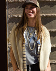 A young woman wearing a brown Ping Pong Surf Club Le Surf Cowgirl Trucker smiles in sunlight, paired with a striped shirt, draped sweater, layered necklaces, and a gray tee while standing against a textured stone wall.