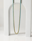 The Faire Turquoise Tennis Necklace, featuring 18K Gold PVD coated stainless steel and turquoise rhinestones, is displayed draped over a white arch-shaped stand on a glossy surface with a soft neutral background.